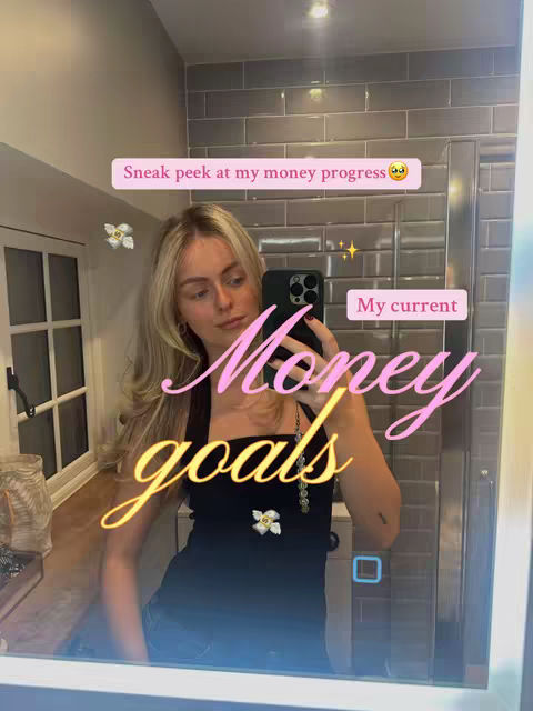 A selfie taken in a bathroom mirror. The person is a woman with blonde hair, wearing a black top. The mirror reflects a tiled wall and a well-lit bathroom. Text overlays include 'Sneak peek at my money progress' and 'My current Money goals', in pink and yellow. The composition is a typical selfie style, with the person holding a phone to capture the image.
