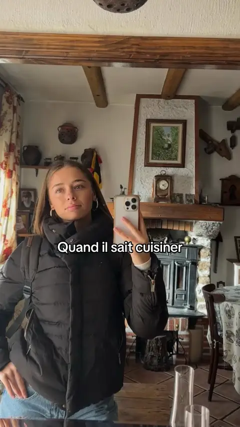 A young woman in a black puffer jacket takes a mirror selfie, her phone obscuring part of her face. She stands in what appears to be a kitchen or dining area with wooden beams, a fireplace, and various decorative items in the background. The text overlay reads 'Quand il sait cuisiner'.