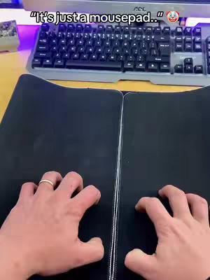 A person's hands are positioned over a black mousepad, the background shows a keyboard and desk setup. The text overlay reads: "It's just a mousepad.." with a laughing emoji.
