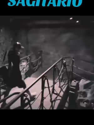 The cover image is a grayscale shot featuring a person walking down a metal staircase or bridge with railings. The architectural elements suggest the scene takes place in a building or a structure with an industrial aesthetic. The person is wearing a long piece of dark clothing. At the top left corner in a bold, teal-colored font, is the word 'SAGITARIO'.