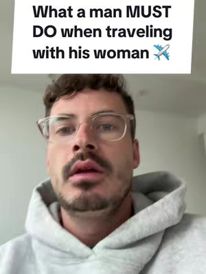 The cover image is a selfie-style shot featuring a man. Above him is a rectangular white banner with black text that reads 'What a man MUST DO when traveling with his woman' along with an airplane emoji. The man is wearing glasses and a grey hoodie. His facial expression is neutral, looking slightly upwards.