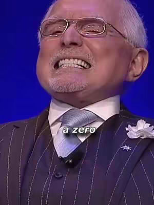 The man is wearing glasses, a suit with pinstripes and a tie. There is a text overlay that says 'a zero'. The background is blue.