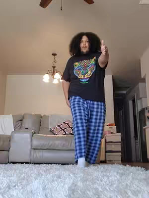 A person stands in a brightly lit living room, pointing their hand out towards the camera. They are wearing a black t-shirt with a colorful design and blue plaid pants. The background features a couch, rug, and a ceiling fan.