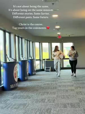 An interior shot shows two women walking through a brightly lit space with large windows. The left side of the frame shows tall blue cylindrical stands with text, while text overlays the image, and the women appear to be chatting. Overall composition is bright and airy, with natural light.