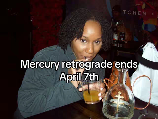A woman with locs is seated at a table, holding a straw in her mouth and looking towards her right. A transparent glass filled with a light-colored beverage is in her hand. The hook text 'Mercury retrograde ends April 7th' is overlaid on the image. Behind her is a blurred bar scene with bottles and a logo.