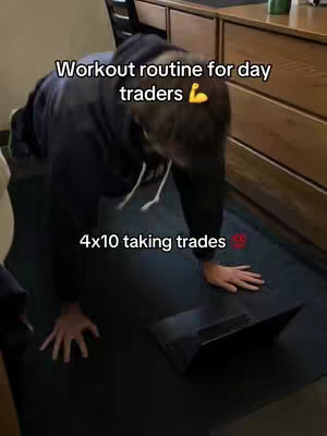 Man is in a push-up position on a black mat, with a laptop open in front of him and text overlays. The background is a wooden desk and wall. The text says 'Workout routine for day traders' and '4x10 taking trades'.