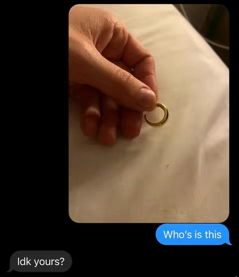 The image shows a hand holding a gold ring, possibly a wedding ring or a similar piece of jewelry. The hand is positioned in the center against a light-colored background that appears to be a bed sheet or similar fabric. In the lower right corner, a blue chat bubble displays the text 'Who's is this?', and in the lower left, a black chat bubble says, 'Idk yours?'. The background is black.