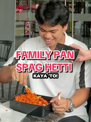 A young man is smiling and holding a tray of spaghetti with tongs. A phone is being held up near his face, displaying an on-screen text overlay. The man is wearing a white shirt. The background is blurred, suggesting a casual restaurant or eatery setting.
