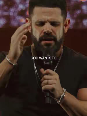 A medium shot captures a man with a short beard and dark hair. He is wearing a black t-shirt and silver bracelets. His left hand is raised near his head, and his right hand holds a microphone close to his mouth. Text that reads "GOD WANTS TO" is overlaid in white at the bottom center.