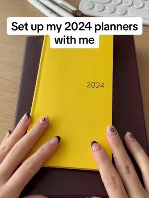 A close-up shot of a person's hands with manicured nails holding a yellow planner labeled "2024". The planner is resting on a dark brown surface. In the background, a keyboard and pens are visible.