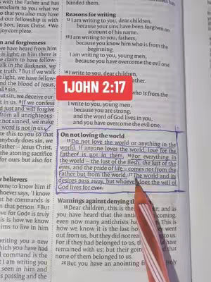 A close-up shot of an open Bible, focused on a highlighted passage with a red rectangular sticker that reads "1 JOHN 2:17". An orange pen is pointing at text in the book suggesting emphasis.