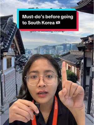 A woman with glasses is holding a small black microphone and pointing up with her right index finger. She's in front of a background that transitions from traditional Korean architecture in the foreground to a modern cityscape.