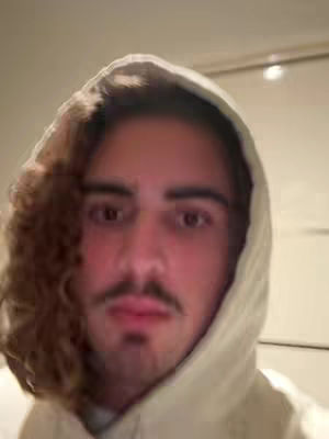 A close-up shot features a young man in a white hoodie. His face is centered, with brown curly hair visible on the left side of his head, framing part of his face. He has a mustache and a slight beard, looking directly at the camera.