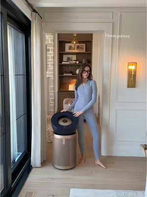 A woman stands barefoot in a well-lit room, facing the camera. She wears a light blue jumpsuit and eyeglasses, holding a Dyson air purifier. The purifier is a cylindrical shape with a black top and a gold base.