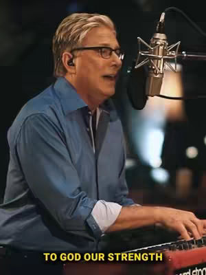 The image shows a man in a blue shirt playing the keyboard and singing into a microphone with text overlayed near the bottom that says 'TO GOD OUR STRENGTH'. The background is blurred, suggesting a concert or studio setting. The man is wearing glasses and an earpiece.