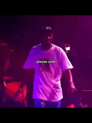 A man wearing a white t-shirt with a graphic is standing on a stage with a microphone in his right hand surrounded by bright purple and red stage lights. The text overlay at the center of his shirt reads 'pisces core'