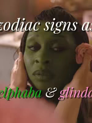 A person with green skin, possibly makeup, is the central focus, with their hands near their ears. Overlayed text labels them as 'zodiac signs as elphaba & glinda'.