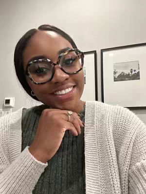A selfie featuring a smiling Black woman with large, patterned eyeglasses. She is wearing a green shirt and a cream-colored cardigan. Her hand is gently touching her cheek, and she is wearing a ring on her finger. Two framed pictures are visible in the background on a white wall. The lighting is good, and the overall image is bright and friendly.