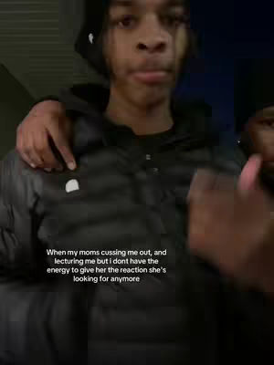 A medium shot shows a young man with dark skin facing the camera. He's wearing a black beanie and a black puffy jacket, and his shoulders are being embraced by someone off-camera. The background is dark with a hint of an urban environment. Text in white overlaid at the bottom of the frame describes a frustrated reaction to a parent, suggesting a youthful tension.