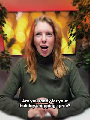 A woman with long, wavy red hair is the central focus, her face framed by a dark green turtleneck sweater. She poses with an open mouth, as if mid-speech, against a softly blurred background of warm, yellow and orange tones suggesting a cozy or festive setting with green plants on each side of her.  The image is well-lit, with a neutral but warm color palette.