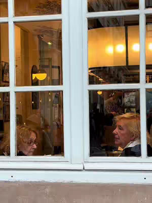 The image shows two women framed by the glass panes of a window. The window has a white frame. The woman visible on the left is wearing glasses and looking out of the window. The woman on the right has blonde hair and is also looking out of the window. The background displays features of an indoor setting, including lights.