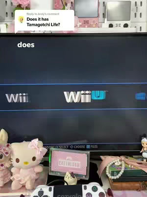 A video game setup is displayed, featuring a dark screen displaying the Wii U interface. In the foreground, a Hello Kitty stuffed toy with a pink bow sits beside a Playstation controller and a couple of other decorative items. The setup features a pink aesthetic with colorful accents from the decorative items.