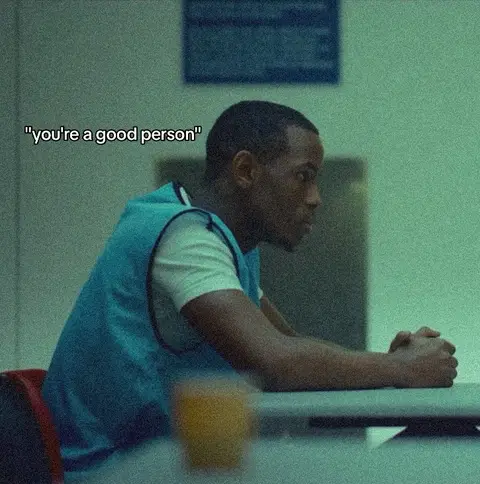 A medium shot shows a Black man seated with his hands clasped in front of him, facing left, with a quote overlayed. He wears a white t-shirt under a blue sleeveless vest. The background suggests an institutional setting with a hint of a window.