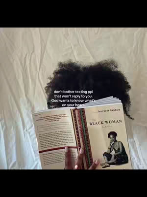 The cover image shows a woman with dark curly hair partially obscuring her face. She's lying on a bed covered with white sheets. She is holding a book open, its title, 'THE BLACK WOMAN', is clearly visible. Above the book, text overlays are present.