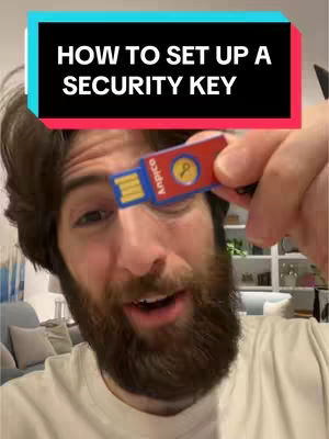 A man holding a USB security key is captured in a selfie-style image. The man, with a full beard and light complexion, is smiling while looking at the camera, with an open and friendly expression. He holds the red and blue security key close to the camera, centered in the frame. The background shows the interior of a home with a couch and what appears to be a bookshelf. A text overlay at the top reads, HOW TO SET UP A SECURITY KEY.