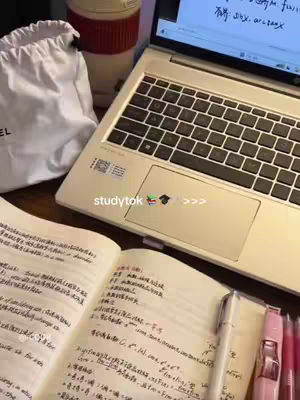 A top-down shot displaying a study setup on a wooden surface. The scene is filled with study materials: an open notebook with handwritten notes, a laptop, pens, markers, a bag, and a covered coffee mug. The lighting is soft, suggesting a well-lit room, and the overall composition is tidy, giving a sense of a focused study environment.