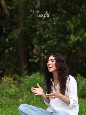 A woman with long, wavy dark hair is seated in a grassy area with trees in the background. The top is white, and she's wearing blue jeans. She is looking upwards with her mouth open as if singing or speaking with passion. The word "JESUS" is written in white at the top of the image.