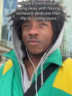 A medium shot shows a Black man looking slightly off-camera with an expression of mild consternation. He wears a gray hoodie under a vibrant, color-blocked green and yellow jacket. White wired earbuds are visible, and the background suggests an urban environment with a building. Text overlayed on the image conveys a message about difficult friendships.
