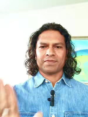 A man with long, wavy black hair and a blue denim shirt is centered in the frame. The background is plain white on the upper portion and a partial view of a painting with blue and green hues on the right, and he is holding one hand up. A small microphone attached to his shirt is visible.