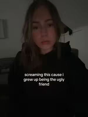A close-up shot of a young woman with light brown hair looking directly at the camera. She is wearing a black hoodie. Overlayed on the image is text that reads "screaming this cause i grew up being the ugly friend". The background is dimly lit, suggesting an indoor environment.