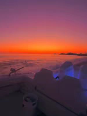 The image captures a scenic overview with a vibrant sunset. The sky transitions from a deep purple at the top to fiery orange and yellow near the horizon. Below, the scene features a layer of mist or clouds, with the suggestion of land formations in the distance. In the foreground, there appear to be some white, or light-colored structures.