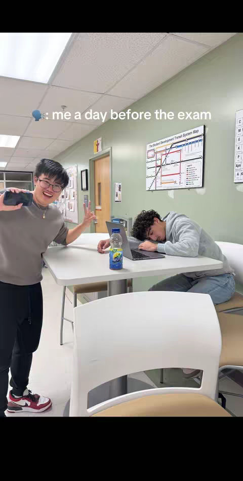 Two young men are pictured indoors. One, standing on the left, is smiling and holding a phone up, making a peace sign. The other, seated at a table on the right, has his head down on his arms as though asleep. A bottle of Nest tea sits on the table. The background includes a wall with a student development transit system map, a door, and a portion of a ceiling.