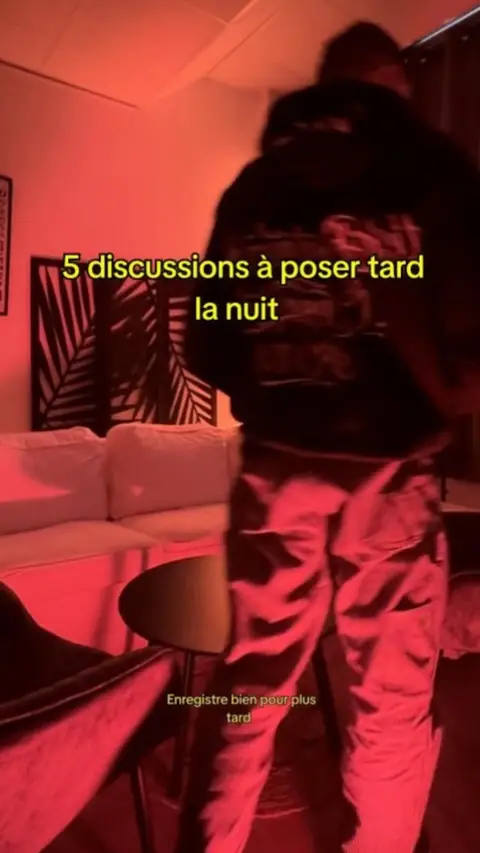 The image is bathed in red light, creating a warm and intimate atmosphere. The frame captures a person, their back towards the viewer, walking with what appears to be another person whose features are obscured due to the dim environment. In the background, an out-of-focus interior scene with a couch, table, and decorative wall panel suggests a living space or a room.