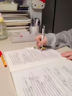 A person's hand is holding a pen and writing on a notebook. The notebook is open, displaying handwritten notes. The setting appears to be a desk with office supplies. There's a Hello Kitty decoration. The image has a warm, well-lit ambiance.
