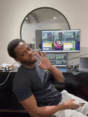 A man is seated in front of a desk with two computer monitors displaying trading charts, holding up a peace sign with his fingers. A large circular mirror is positioned behind him, and various office supplies are visible on the desk. The man is dressed in a dark t-shirt and light colored pants.