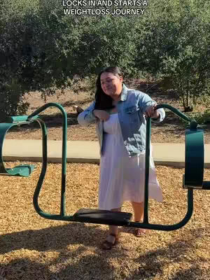 A woman stands confidently in a sunny outdoor setting, leaning on a playground structure. She wears a white dress and a light blue jean jacket, with foliage in the background.