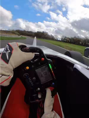 A first-person perspective from inside a race car. The shot captures the driver's gloved hands on the steering wheel, the car's dashboard, and the track ahead with a cloudy sky.