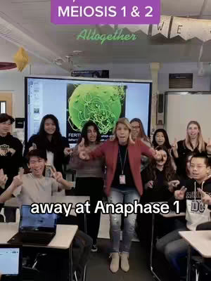 An image of a classroom with a teacher and students posed. The teacher is in the center, gesturing with both hands. Students are seated at desks in front and to the sides of her, some with thumbs up. Behind the teacher, the screen displays the green image of a cell and its components.