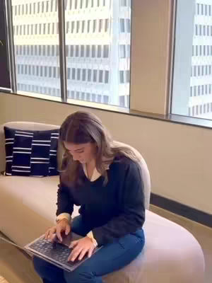 A young woman sits on a beige sofa in front of a window with a city view. She's focused on a laptop, wearing a black sweater and blue jeans. The room has a minimalist design with neutral colors and a decorative pillow with a black and white pattern.