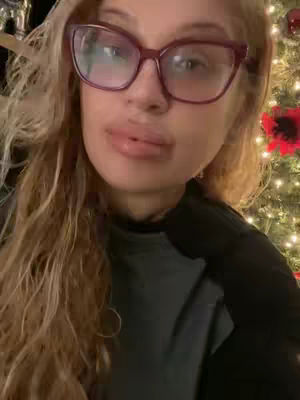 A woman with long blonde hair and large eyeglasses, wearing a black top. She's looking at the camera with her lips slightly parted. Behind her is a blurred Christmas tree with lights and a red ornament.