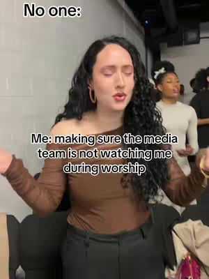 The image captures a woman with long, dark, curly hair standing in what appears to be a backstage or studio setting. She is wearing a brown, off-the-shoulder top and black pants. Her arms are raised, and her eyes are closed, suggesting a moment of worship or performance. A person is slightly visible in the background. Text overlays read "No one:" and "Me: making sure the media team is not watching me during worship."