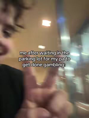 The cover image shows a selfie-style shot with the creator in the foreground, focusing on their face and hand gestures. The background appears blurry and indistinct, possibly depicting a dimly lit indoor environment. The text overlay, in a clear font, describes a relatable situation: waiting in a parking lot while someone is gambling.