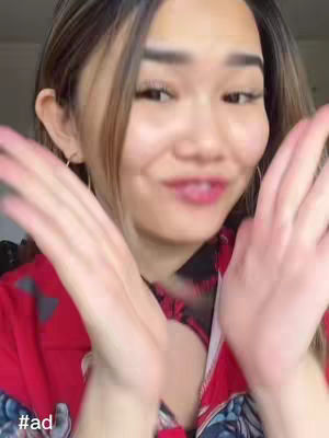 A selfie-style video cover image features a young woman with light skin and long, light brown hair. She stands against a blurred background. The woman is wearing a red top and has her hands up near her face, as if in a gesture of surprise or excitement. The image is well-lit, with a soft and flattering glow on the woman's face. The text overlay '#ad' is positioned at the bottom left corner.
