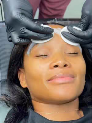 A close-up shot of a Black woman's face while someone is applying a skincare treatment, such as wiping product with cotton pads. The woman is positioned in the center of the frame with her eyes closed, suggesting a relaxing or beauty treatment experience. The background is blurred, indicating focus on the face. Hands in black gloves are seen performing the treatment.
