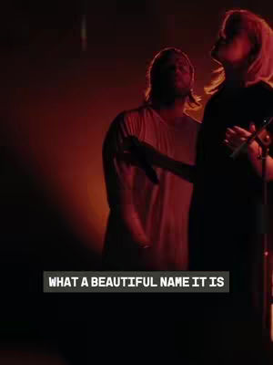 The image shows two people on a stage with dim red lighting, presenting what could be a musical performance. The person on the right is centered, with a microphone in hand and the other person's figure is partially visible behind them. The text overlayed reads, "WHAT A BEAUTIFUL NAME IT IS". The overall composition focuses on the performers with a soft, warm lighting.