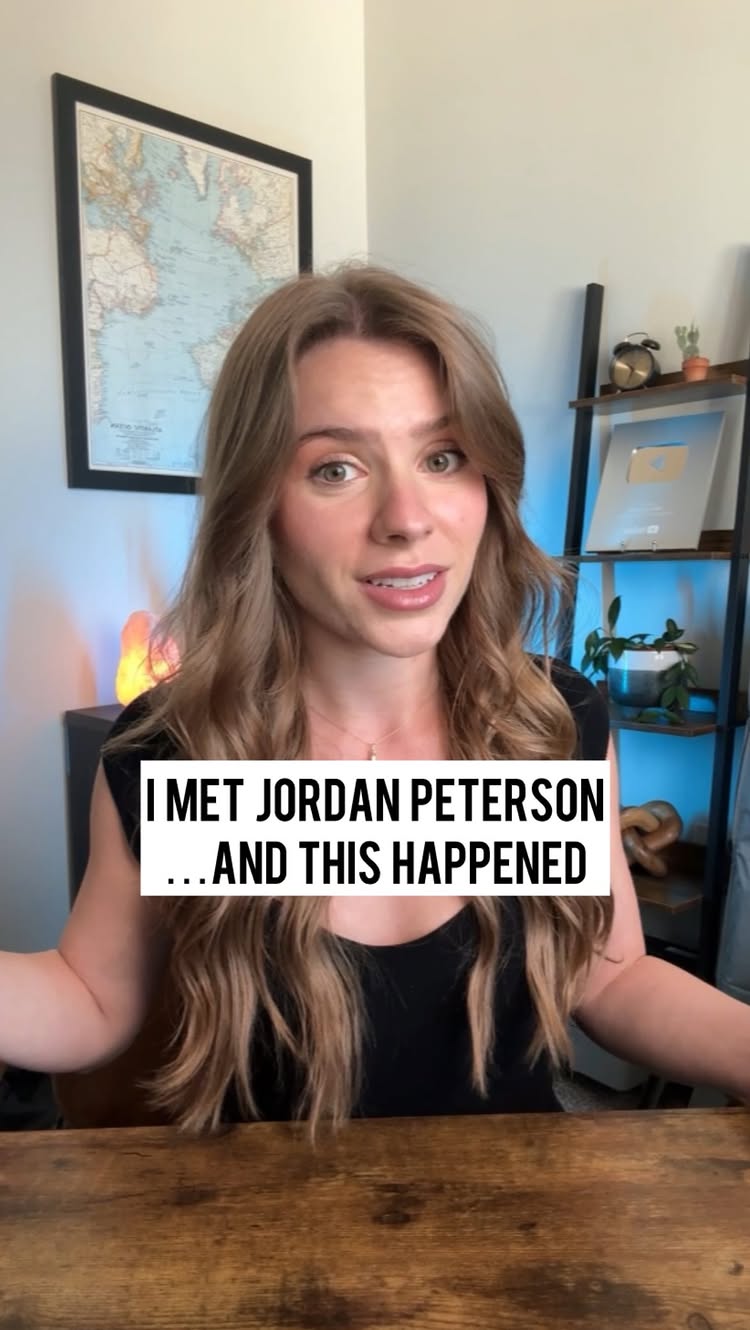 A woman with long wavy blonde hair is centered in the frame, smiling and speaking directly to the camera. She is wearing a black top. Behind her is a wooden desk, a map, and some shelves with items on them, suggesting a home office or studio environment. A text overlay at the bottom reads, 'I MET JORDAN PETERSON...AND THIS HAPPENED'. The background features soft lighting.